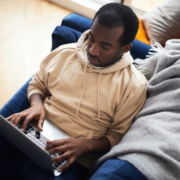 Young Man Relaxing At Home Lying On Sofa Using Laptop To Work From Home Or Shop Online